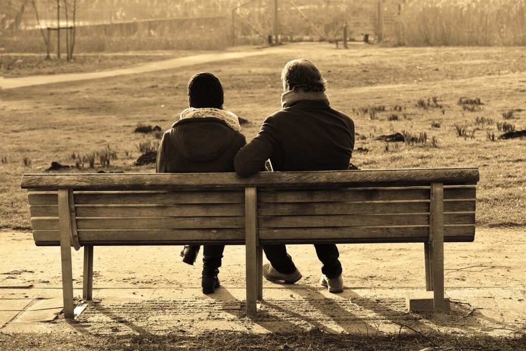 Zwei Menschen, die auf einer Bank sitzen. Man sieht sie von hinten und sie schauen in die ferne Landschaft.