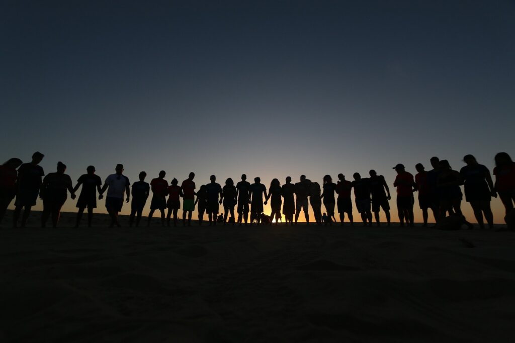 Als Symbol für das Netzwerktreffen stehen am Horizont viele Menschen nebeneinander. Im Hintergrund geht die Sonne unter.
