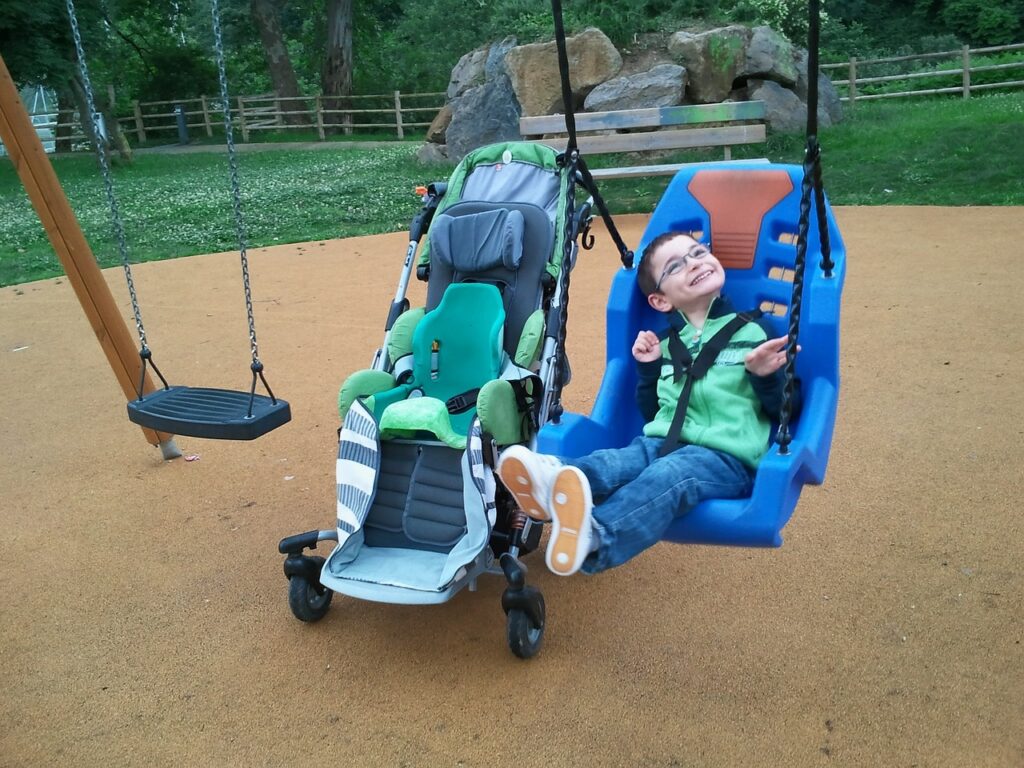 Kind mit Behinderung, welches in einer Schaukel sitzt und sich freut. Daneben steht sein Rollstuhl/Buggy.